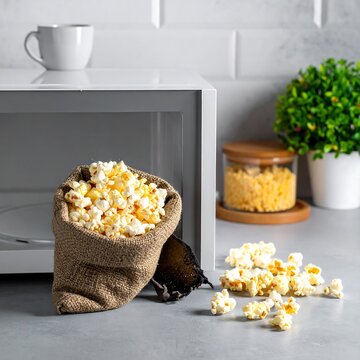 A rustic burlap bag overflows with fresh popcorn next to an open microwave in a bright, modern kitchen setting