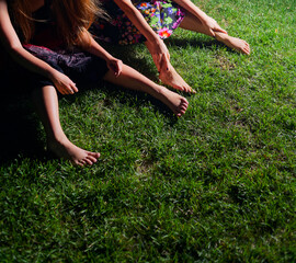 Relaxation Outside: Young Women Sitting on Lawn in Colourful Dresses Engaging in Leisurely Summertime Night Gathering, Focus on Legs and Bare Feet Copyspace