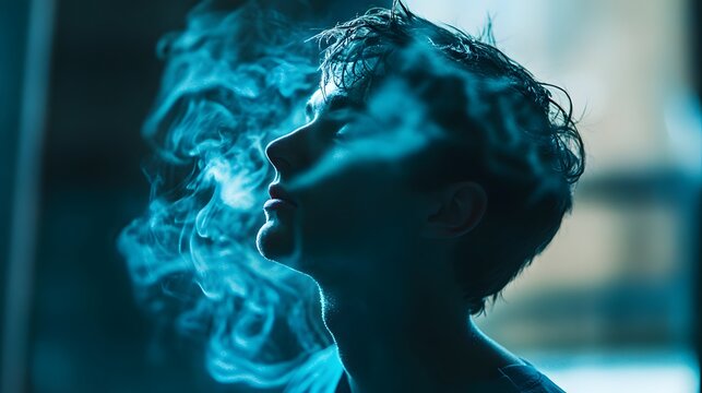 A young man exhales a cloud of blue smoke, creating a mysterious and artistic portrait. His face is partially obscured by the smoke, adding to the sense of intrigue.