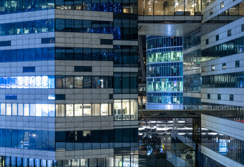 Tall buildings show bright windows at night. Some offices are active with lights on, while others are dark. The design features glass and concrete structures.