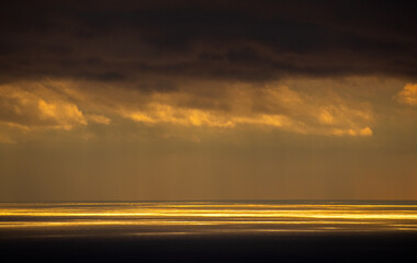 Clouds cover the horizon as sunlight reflects on the ocean in the early evening sky near the coast. Dark clouds hang above, creating a contrast with the bright reflection below.