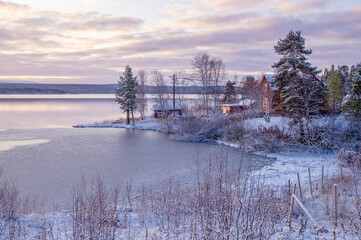 A rural scene shows a frozen lake with patches of ice. Houses stand next to the water as trees surround the area. The sky has clouds with colors from the sunset.