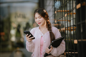 Fototapeta premium A young woman stands outdoors holding a smartphone and laptop, smiling confidently in a modern urban setting, representing a mobile lifestyle, technology use, and smart working concept.