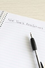 Close up shot of handwriting "New Year's Resolutions" on new notebook with black pen on white wooden table.