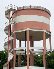 Elevated water storage tank with blue railings on stilts, standing against green trees and cloudy sky, a common rural water supply infrastructure.