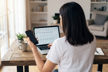 Rear view of woman working on laptop and holding mobile phone at wooden desk in bright home office