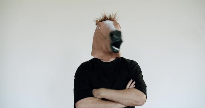 A man wearing a horse head mask stands with his arms crossed. He looks around, observing his surroundings. His posture suggests confidence and a sense of humor.