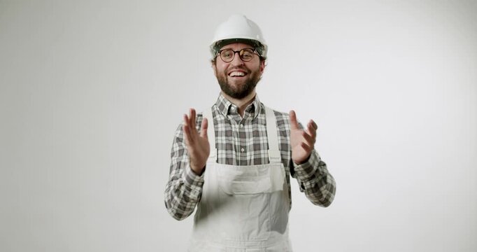 A cheerful builder in white overalls and a hard hat claps his hands and smiles at the camera. He is happy with the progress and results of his work. The perfect shot for your construction