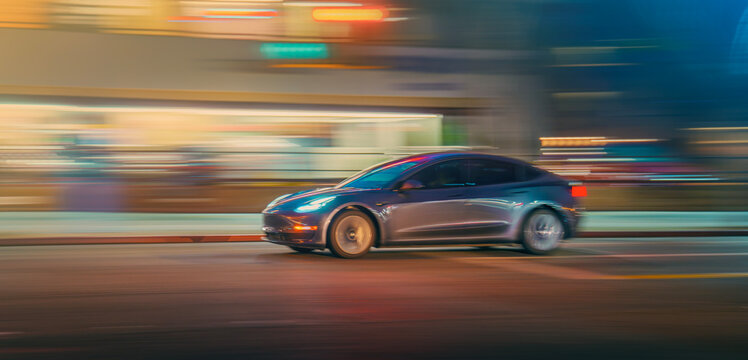 LOS ANGELES, CA - December 25, 2025: Tesla Model 3 electric car driving by a colorful neon illumination on a city street at night. Blurred motion
