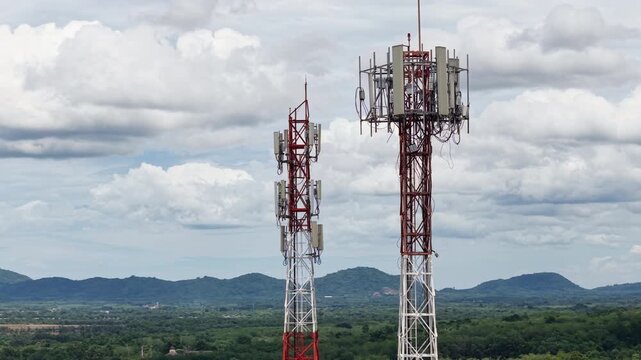 Aerial 4K cinematic view of telecommunication towers for 5G cellular network against a scenic mountain landscape and cloudy sky.