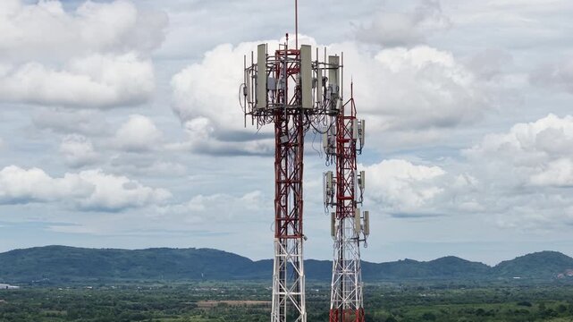 Aerial 4K cinematic view of telecommunication towers for 5G cellular network against a scenic mountain landscape and cloudy sky.