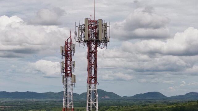 Aerial 4K cinematic view of telecommunication towers for 5G cellular network against a scenic mountain landscape and cloudy sky.