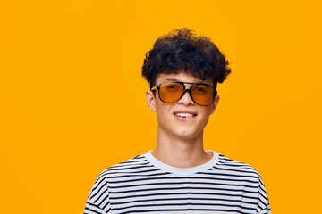 Smiling young man wearing orange sunglasses and striped shirt stands against a bright yellow background, radiating cheerful and trendy vibes with casual hairstyle. © SHOTPRIME STUDIO
