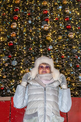 Mature adult female in winter clothes on the background of Christmas tree in old town square with night illumination during of snow fall.