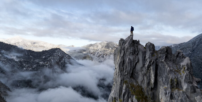 Person Standing On Jagged Peak Above Snowy Mountains With Cloudy Valley View - Powered by Adobe