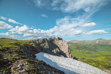 Sunlit grassy meadow, glacier and rocky cliff on precipice edge above alpine valley against big hills under clouds in blue sky in sunny day. Scenic view with green grass near sharp rocks in bright sun
