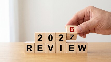 A hand carefully positions a wooden block with the number six onto a stack spelling out 2027 review symbolizing assessment