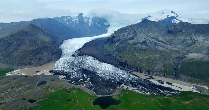 Aerial view of a valley glacier spilling into a muddy meltwater lagoon, surrounded by rugged mountains and black volcanic plains under cloudy summer skies.