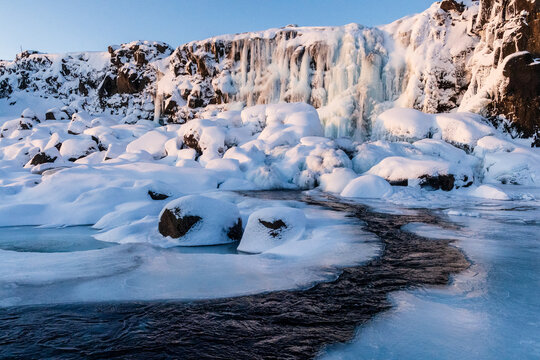 Winter outdoor scene showing a snow-covered river near the partially frozen Oxararfoss Waterfall in Thingvellir national park in the Golden circle in Iceland.