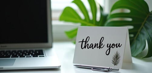 White card with thank you text and pen sit near laptop on white office desk. Green plant adds natural touch to workspace. Simple expression of appreciation.