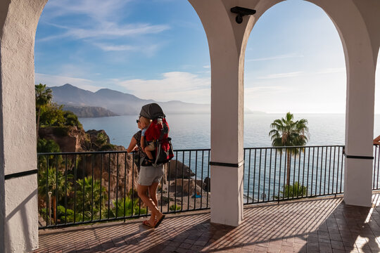 A mother carrying her baby in a backpack stands beneath the white arches of Balcon de Europa, admiring the panoramic coastal cliffs and sea views in the sunny destination of Nerja in southern Spain.