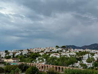 Obraz premium Historic Eagle Aqueduct with brick arches spans across a lush green valley, standing below white hillside villas and dramatic mountains under a cloudy sky near the coastal town of Nerja, Spain.