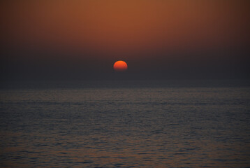 A stunning sunset over the calm sea horizon with a vibrant red sun setting into the misty water. The deep orange and dark blue sky creates a peaceful, minimalist atmosphere. 