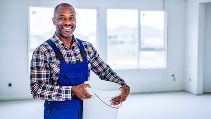 Smiling black builder holding white bucket in empty room with windows  