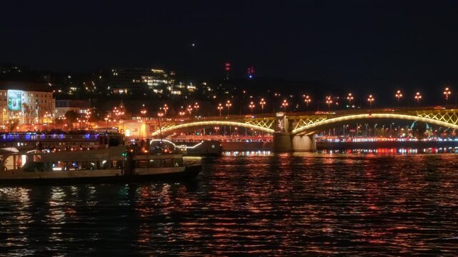 Panoramic view of Budapest city at night, architecture with warm golden illumination reflecting in Danube river, featuring passing cruise boats, and Margit bridge view