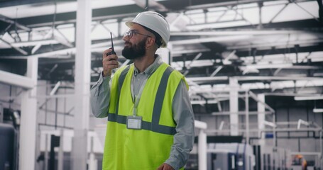Engineer Communicating Via Walkie Talkie in Factory. Bearded Engineer in Hard Hat and Safety Vest Uses Walkie Talkie Coordinate Teams on Large Industrial Site. Operational Report, Project Management.