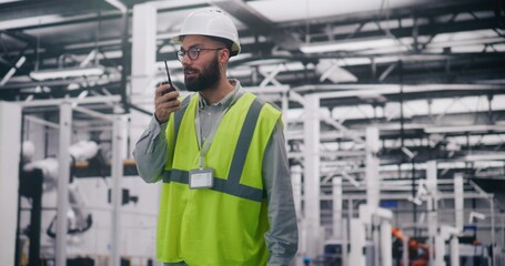 Engineer Communicating Via Walkie Talkie in Factory. Bearded Engineer in Hard Hat and Safety Vest Uses Walkie Talkie Coordinate Teams on Large Industrial Site. Operational Report, Project Management.