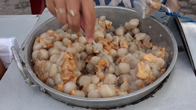 Street food vendor serving siomay and cilok, stirring hot dumplings while assisting customers at a local stall. Authentic Indonesian street food scene with fresh, homemade snacks.