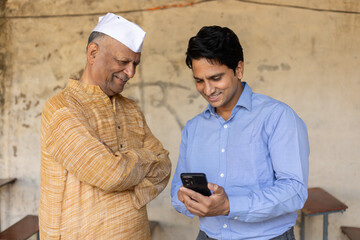 Fototapeta premium Young indian bank person or finance adviser showing information to senior farmer on smart phone. Government scheme, Agriculture or business loan, Rural india, Support and assistance.