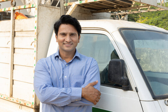 Confident young Indian man driver standing with his arms crossed next to a white mini truck, small business ownership and delivery services. Transport.