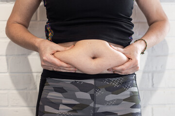 A woman holds a fold of subcutaneous fat protruding from under her sports shirt with both hands against a white wall, demonstrating the extent of her obesity. Obesity and fitness