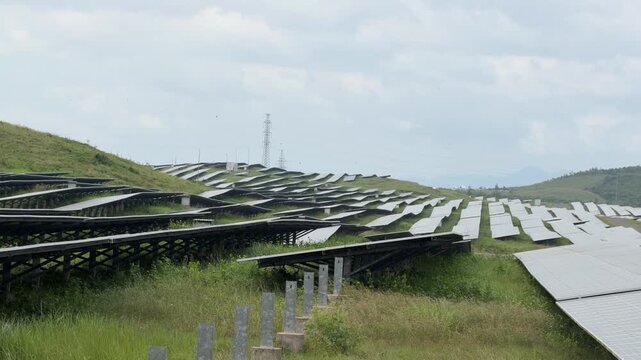 Solar panel grids stretch across grassy land while overhead clouds drift slowly, illuminating parts of the large renewable landscape. Green technology.