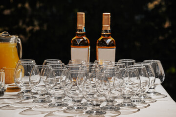 Row of empty cognac snifter glasses arranged on a white table with two bottles of beverage and a pitcher of juice, preparing for a festive event or celebration