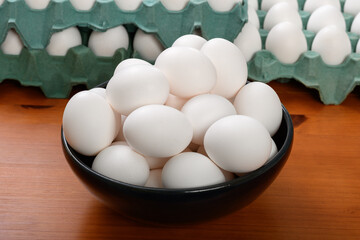 Fresh white eggs stacked in a black bowl with egg cartons at the bottom.