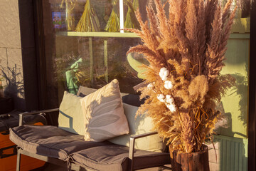 A bench with soft cushions and a vase of reed branches in front of the cafe window. An outdoor seating area.