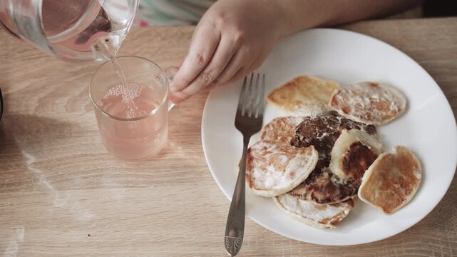Unrecognizable person pours compote into mug from pitcher during breakfast with pancakes, top view. Woman pours fortified, homemade, fruity, authentic drink to replenish fluids.