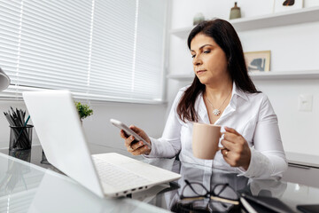 A professional woman in a white shirt sits at a desk, multitasking with a smartphone and a cup of coffee while working on a laptop in a bright office