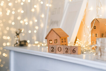 Wooden house models and 2026 number blocks on a white surface with blurred festive lights in the background. New Year decoration for home interior.
