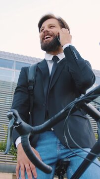 Stylish man in black jacket and jeanse talking with interlocutor on smartphone and laughing while stopped on his bicycle near beautiful city building