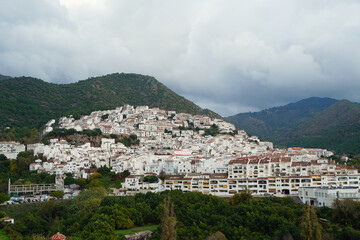 Elevated view of the whitewashed hillside town of Oj&eacute;n in Andalusia, with dense traditional houses framed by green mountains and a cloudy sky, highlighting its historic Mediterranean setting