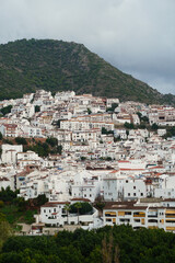 Elevated view of the whitewashed hillside town of Oj&eacute;n in Andalusia, with dense traditional houses framed by green mountains and a cloudy sky, highlighting its historic Mediterranean setting