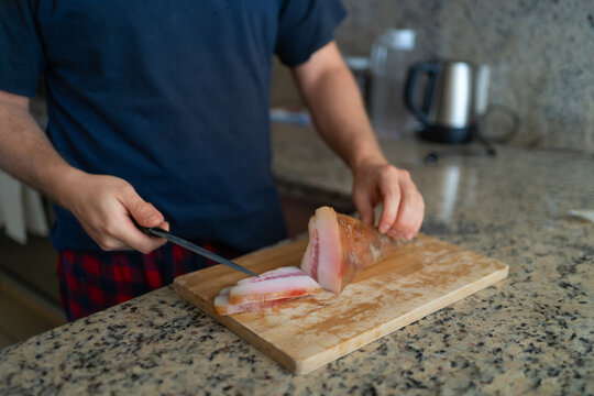 Una persona est&aacute; cortando  un trozo de guanciale en rebanadas con un cuchillo en una tabla de madera.