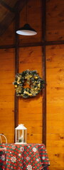 Christmas wreath with yellow and golden decorations on wall of wooden cabin, above table with seasonal tablecloth and lantern with candle
