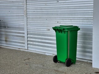 Green wheeled trash bin standing outdoors against metal wall in urban industrial setting
