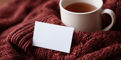 a blank white postcard mockup tucked into the sleeve of a chunky knit burgundy sweater. A steaming mug of hot apple cider is blurred in the background