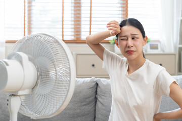 Suffer summer heat wave stroke, hot weather, tired asian young woman sitting on couch in living room without air-conditioner, using electric fan, wave fan to cool for ventilation when high temperature